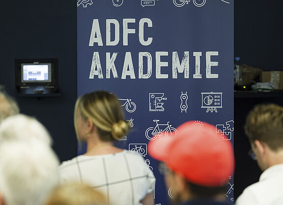 Mehrere Menschen blicken auf eine Bühne. Dort ist ein Banner mit der Aufschrift "ADFC-Akademie" zu sehen.