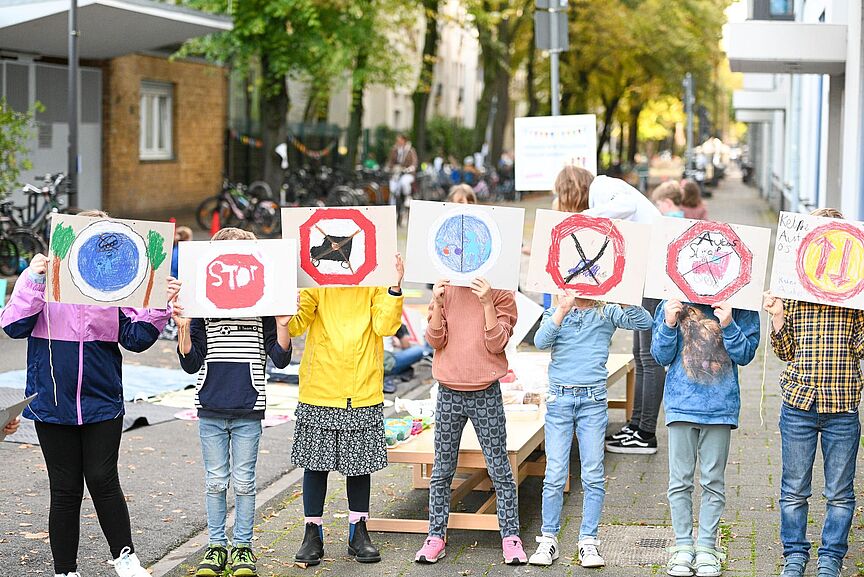 Schulstraßen in NRW Kinder blockieren eine Straße, halten Plakate hoch und setzen sich damit für die Einrichtung von Schulstraßen ein.