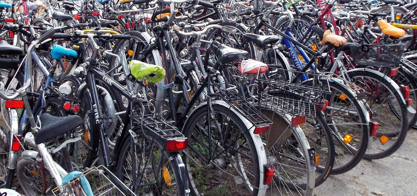 Hunderte von Fahrrädern verschiedener Farben und Modelle stehen dicht gedrängt und ungeordnet auf einer befestigten Fläche; die Räder sind ineinander verschachtelt, mit Körben, Taschen und bunten Sattelbezügen ausgestattet, ohne erkennbare Parkordnung oder Abstellvorrichtungen.