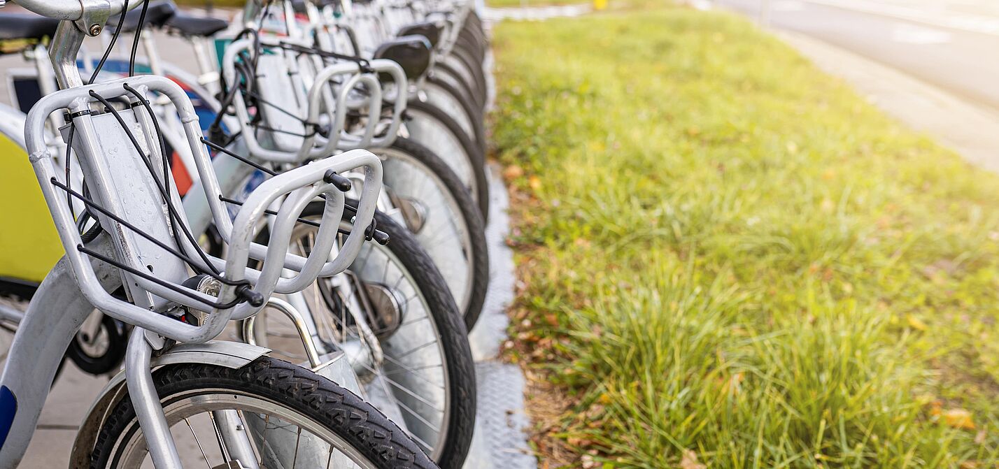 Eine Reihe von öffentlichen Leihfahrrädern mit silbernen Rahmen und grauen Körben am Lenker steht ordentlich nebeneinander an einer stationsgebundenen Verleihstation. Die Fahrräder sind auf einem gepflasterten Gehweg abgestellt, neben dem eine Grünfläche verläuft.