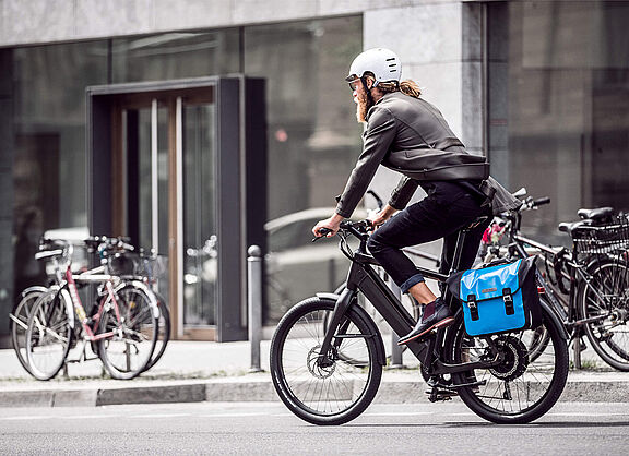 Ein Mann mit Bart und weißem Helm fährt auf einem modernen Elektrofahrrad durch eine städtische Straße. Er trägt Alltagskleidung (Jacke und Jeans) und hat eine leuchtend blaue Packtasche am Gepäckträger befestigt. Im Hintergrund sind Gebäude mit großen Glasfronten und parkende Fahrräder unscharf zu erkennen.