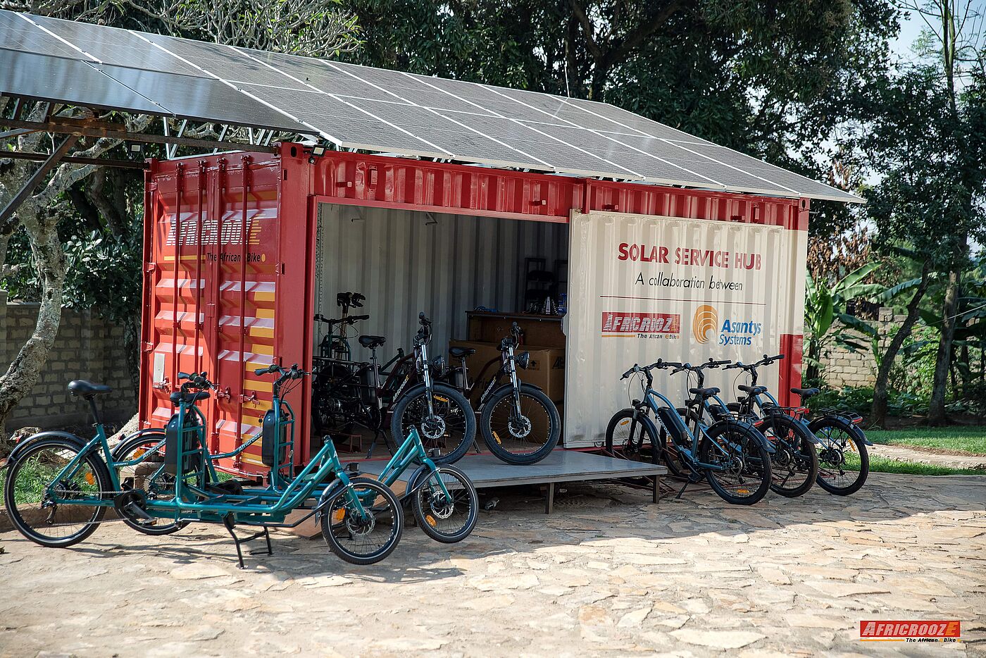Ein Schiffscontainer mit der weißen Aufschrift „Solar Service Hub“ steht im Freien unter einem breiten Dach aus Solarpaneelen. Im geöffneten Container und auf dem gepflasterten Platz davor stehen mehrere schwarze und türkisgrüne Elektrofahrräder sowie Lastenräder.