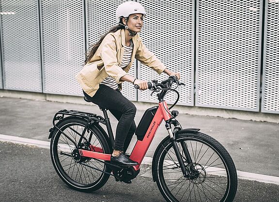 Eine Frau fährt auf einem roten Elektrorad auf Asphalt, im Hintergrund graue, gelochte Metallwände. 